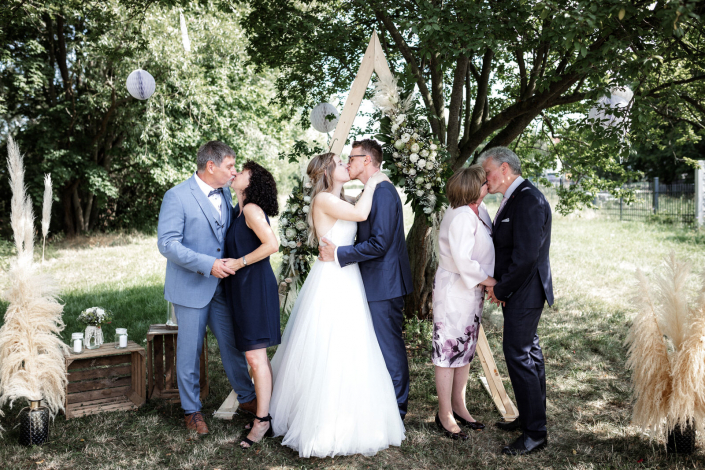 Brautpaar mit Eltern unter vintage Traubogen küssen sich